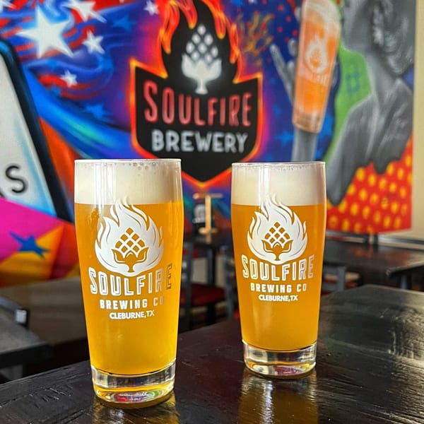 Two Soulfire Brewing Co. pints of golden beer sit on a wooden table before a colorful brewery mural.
