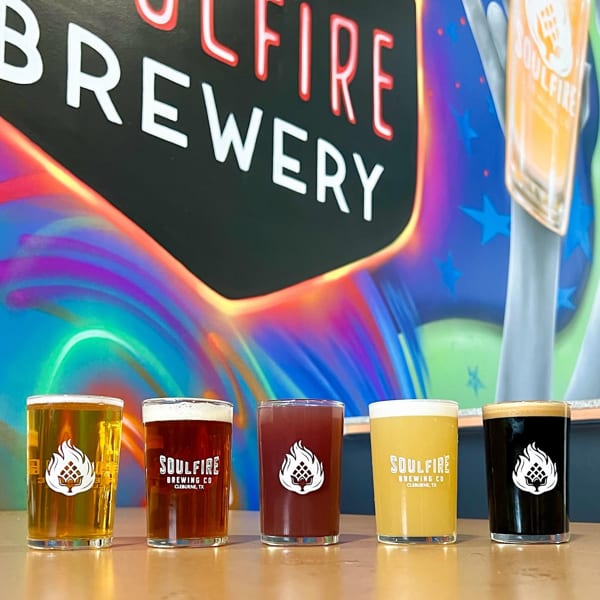 Five Soulfire Brewing Co. beers in varied colors line a wooden table against a vibrant mural.