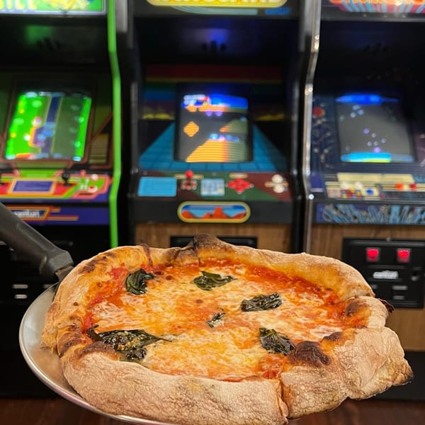 Large cheese pizza with basil and tomato sauce held before colorful arcade machines.