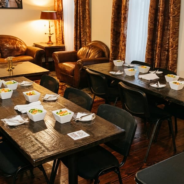 Restaurant dining room with two long wooden tables set with salads, and a lounge area with leather furniture.
