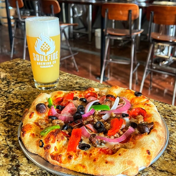Loaded pizza with pepperoni, sausage, olives, peppers, onions, and mushrooms beside a SoulFire Brewing Co. beer on a granite table.